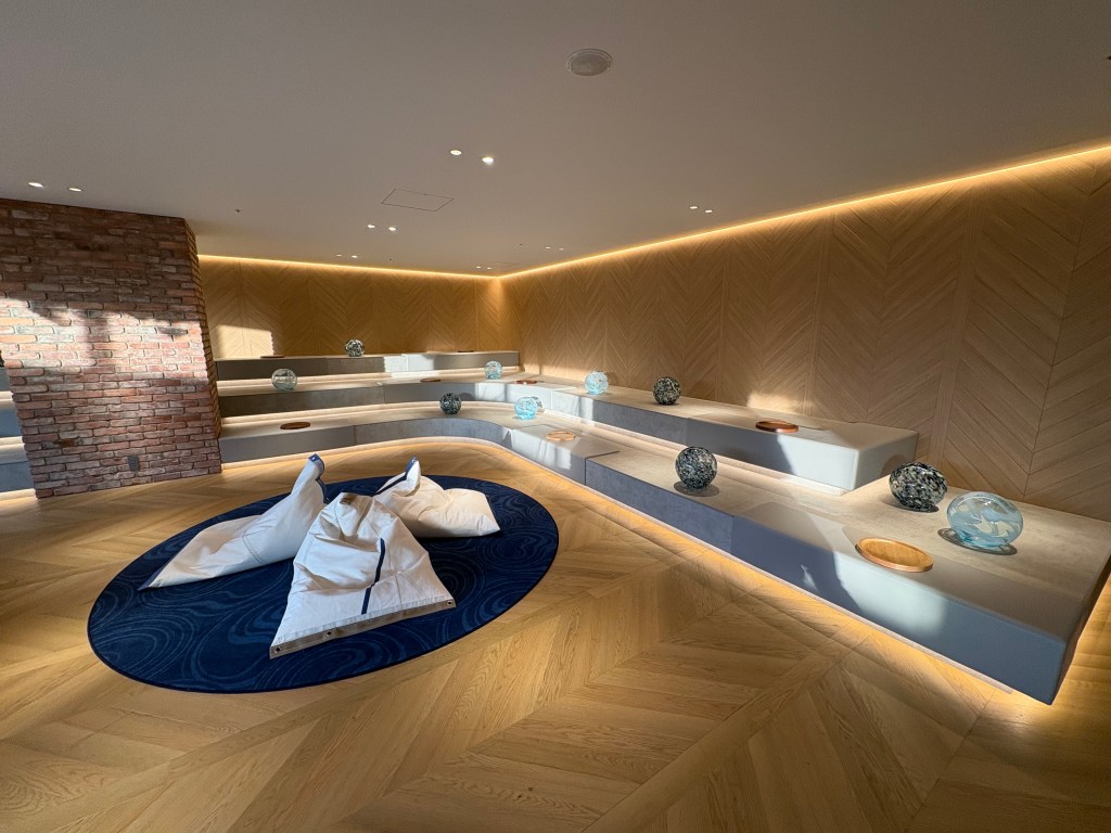 A photograph taken from inside the dimly lit hot spring public bath, looking out through large windows at the complex of train tracks and station platforms.