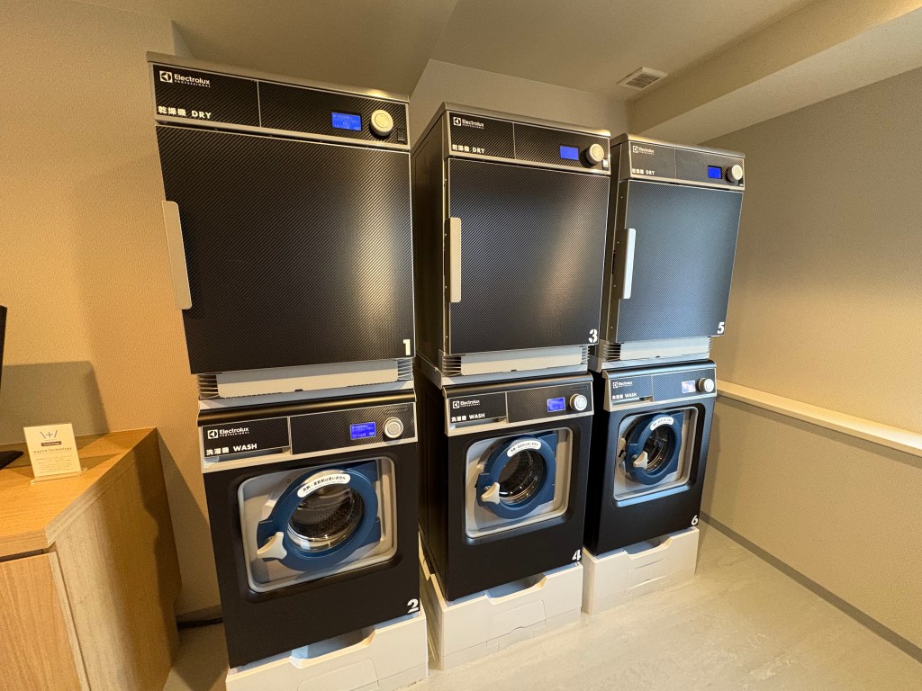 A photograph of the hotel's coin laundry room, featuring a row of clean, modern stainless steel washer and dryer units.