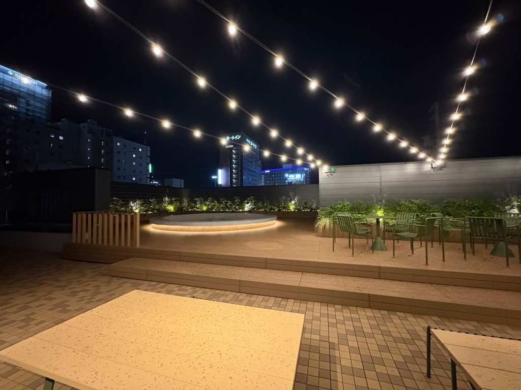 A low-light photograph focusing on a single large glass window in the relaxation area, offering a panoramic view of the sparkling Hakodate cityscape at night.