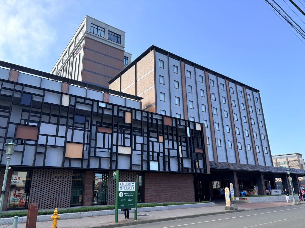 Exterior view of the Hakodate Kokusai Hotel building on a sunny day, showcasing modern geometric facade panels, brick architecture, and street entrance.