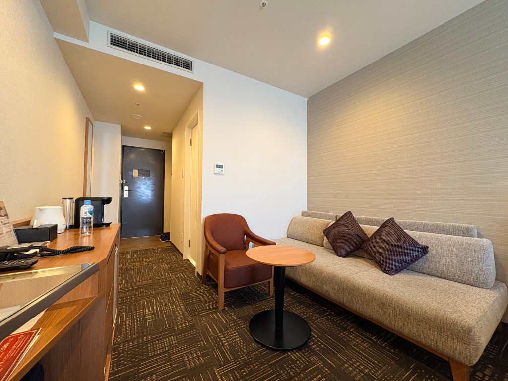 Seating area of the Premium Japanese Twin room, showing a comfortable fabric sofa, side table, armchair, and the hallway leading to the entrance door.