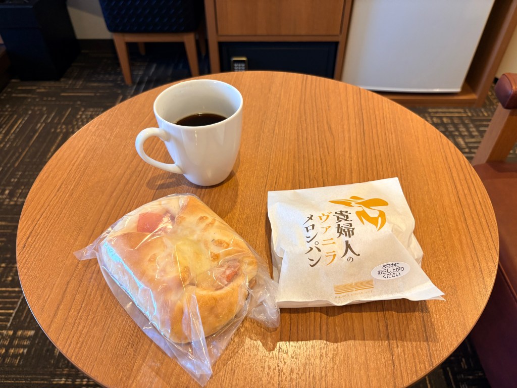 The hotel's packaged vanilla melon pan, a savory cheese bread roll, and a white mug of black coffee placed on a round wooden table in the guest room.