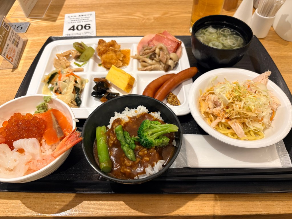 Breakfast buffet tray at Hakodate Kokusai Hotel featuring a custom seafood bowl topped with salmon, ikura, and squid, alongside vegetable curry, noodle salad, miso soup, and partitioned plate of regional sides.